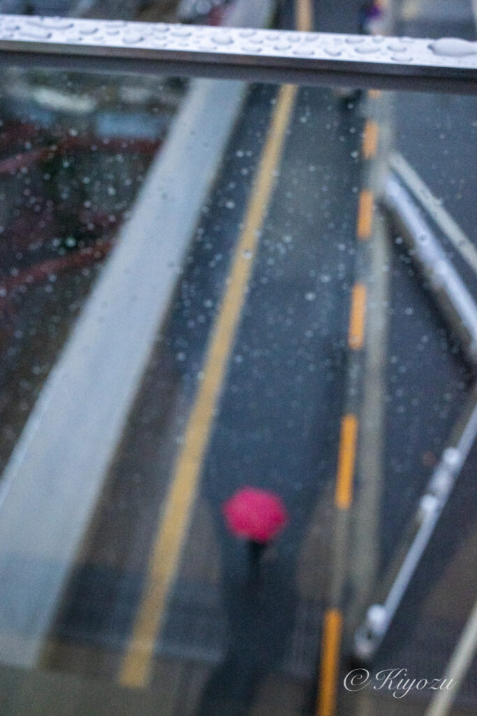 雨粒に濡れた壁越しに撮る、赤い傘の人