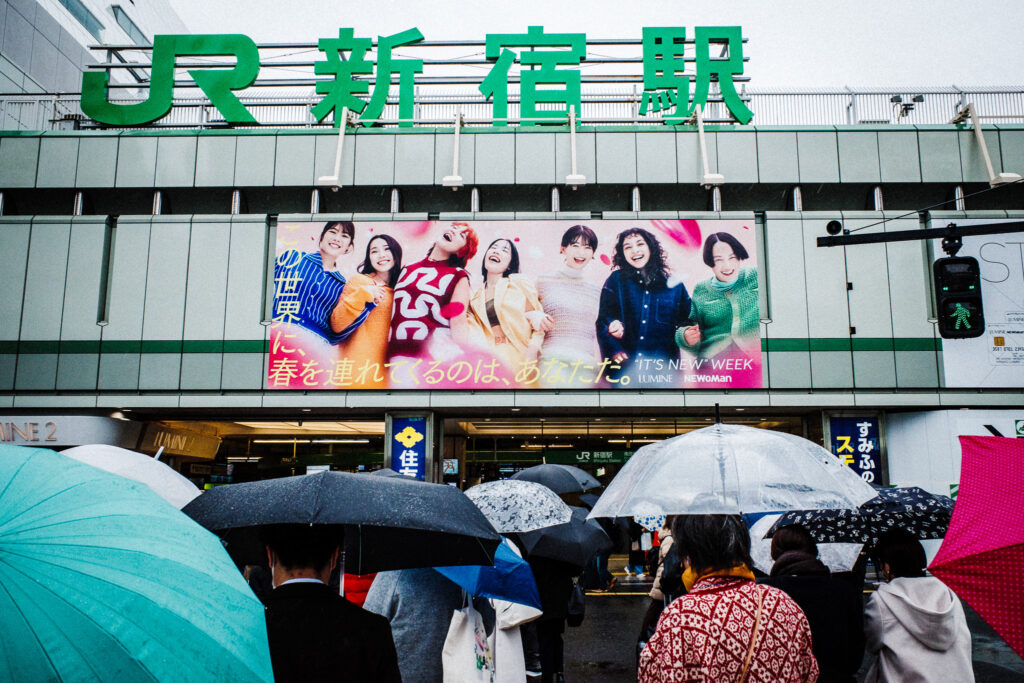 雨の新宿駅前のストリート写真。「明るい表情を浮かべる看板広告の若者と、駅に向かう人々」